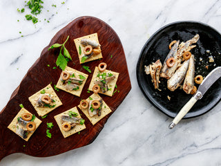 A rustic cutting board serves saltines topped with Patagonia Provisions Spanish White Anchovies, olives and fresh parsley, one a white wooden table beside a skillet of anchovies and a glass of beer