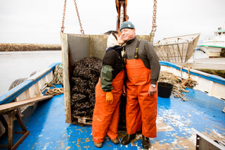 A man and woman on a boat wearing protective overalls and hats and the woman is kissing the man on his cheek