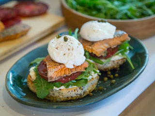 A sliced seed bagel side-by-side on a blue plate topped with greens, large tomato slice, Patagonia Provisions Wild Salmon, and poached egg
