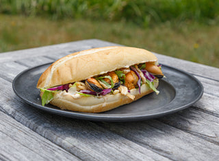 A hoagie with a layer of greens, remoulade spread, and mussels with potato chips on the side on a rectangular plate