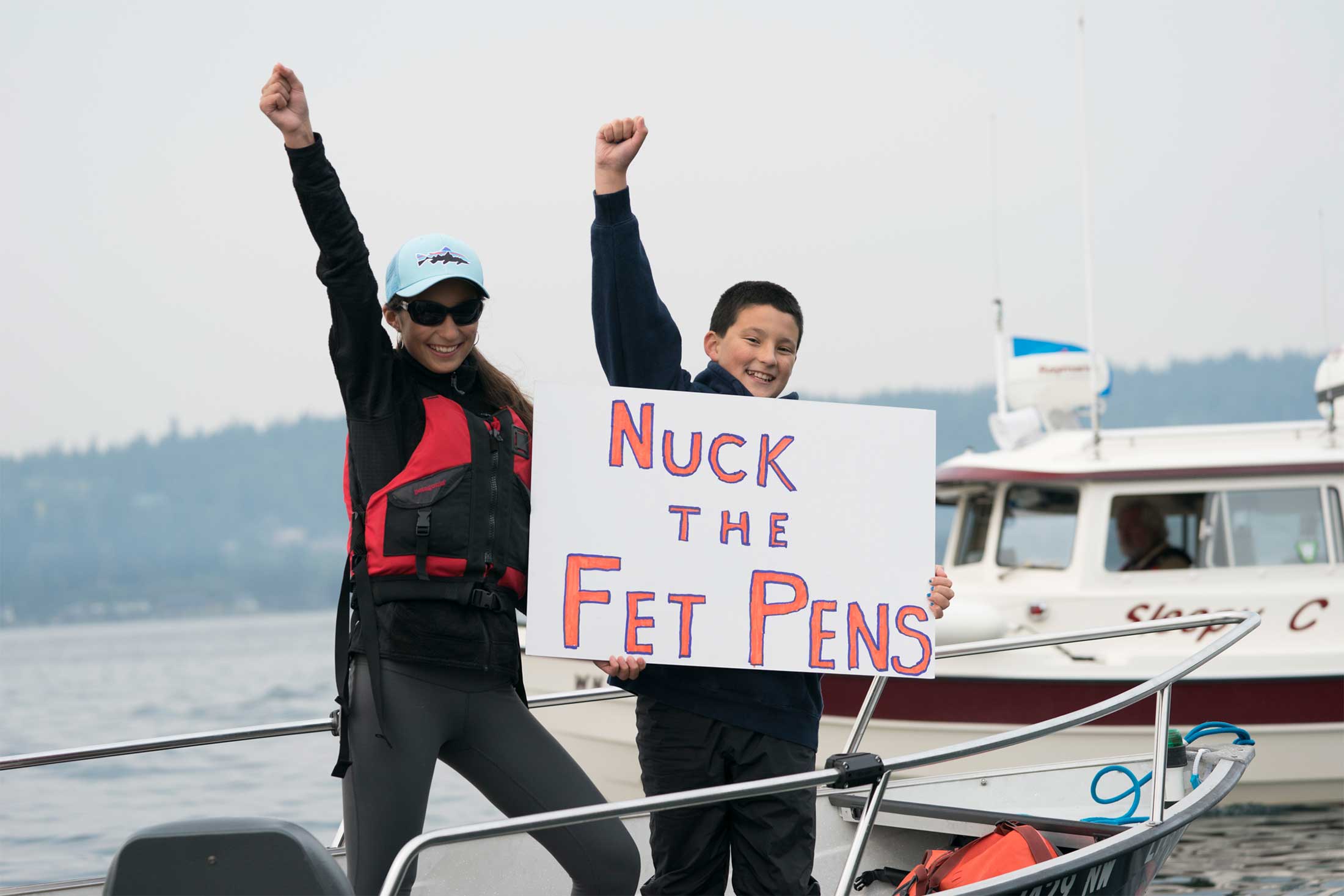 Two young activists stand on a boat, holding a sign and fists in the air, protesting Net Pens in Puget Sound