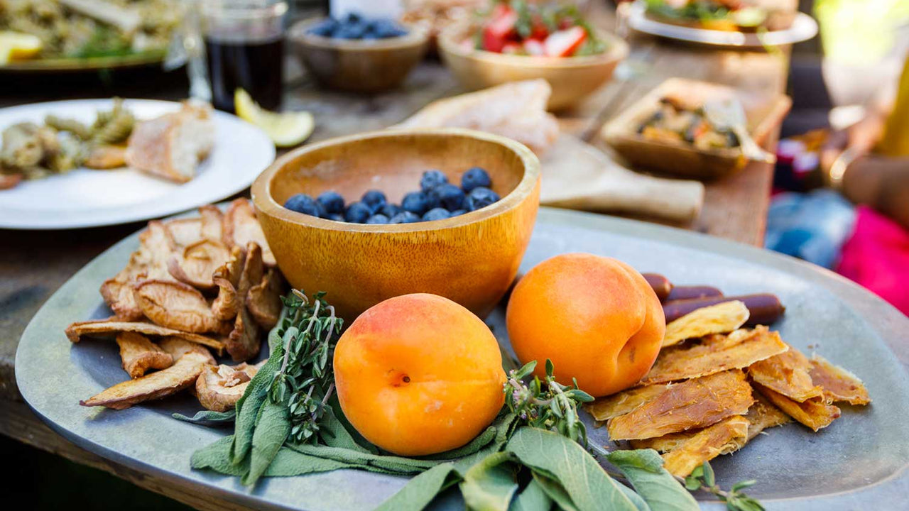 A metal tray topped with fresh and dried fruits and herbs rests at the end of a long wooden table covered with dishes of food and drinks