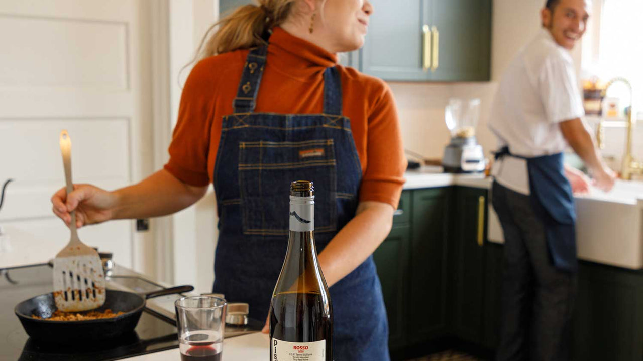 Two home chefs wearing blue denim aprons work together in the kitchen, preparing a meal beside a bottle and glass of Patagonia Provisions wine