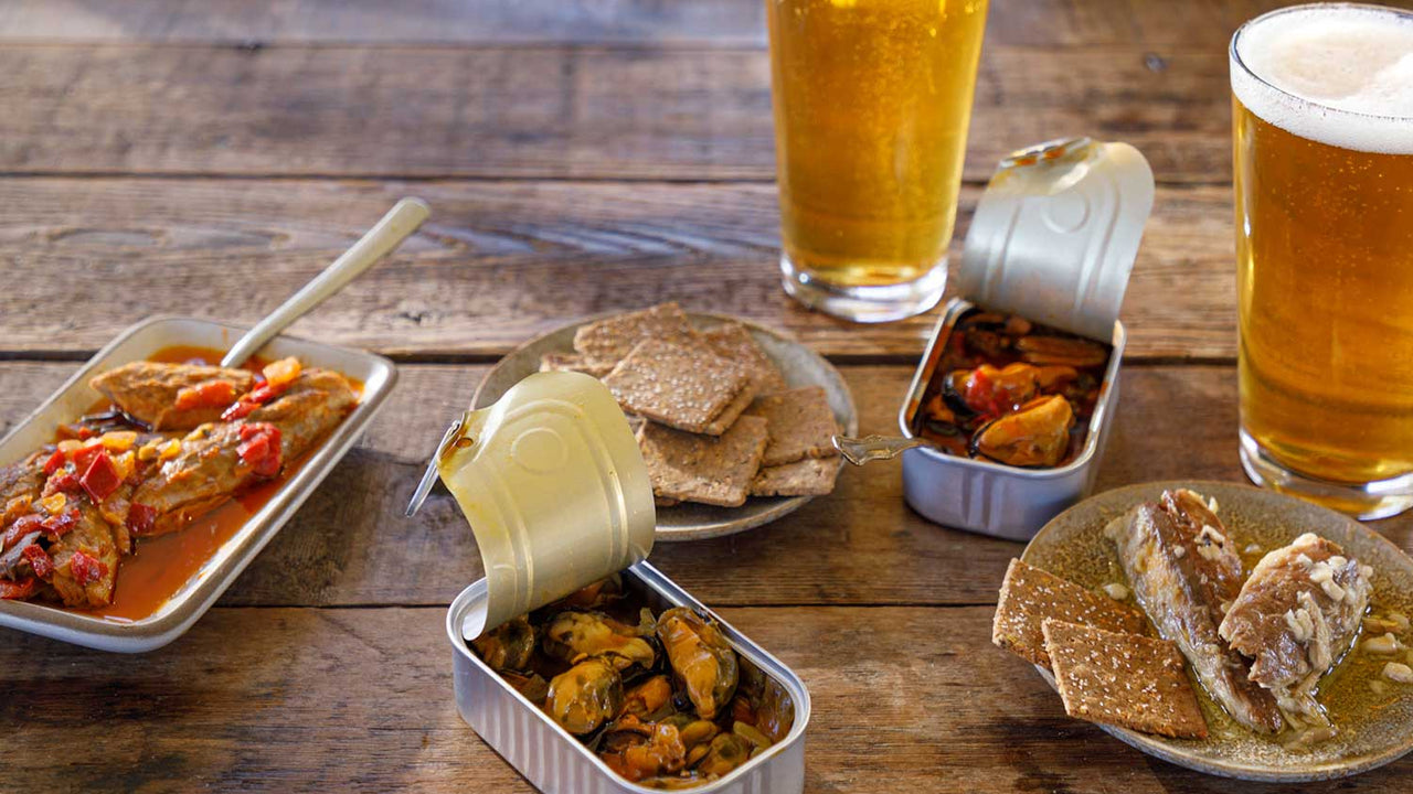 A wooden table is covered with open tins and plates offering Patagonia Provisions seafood, beside crackers and golden pints of beer