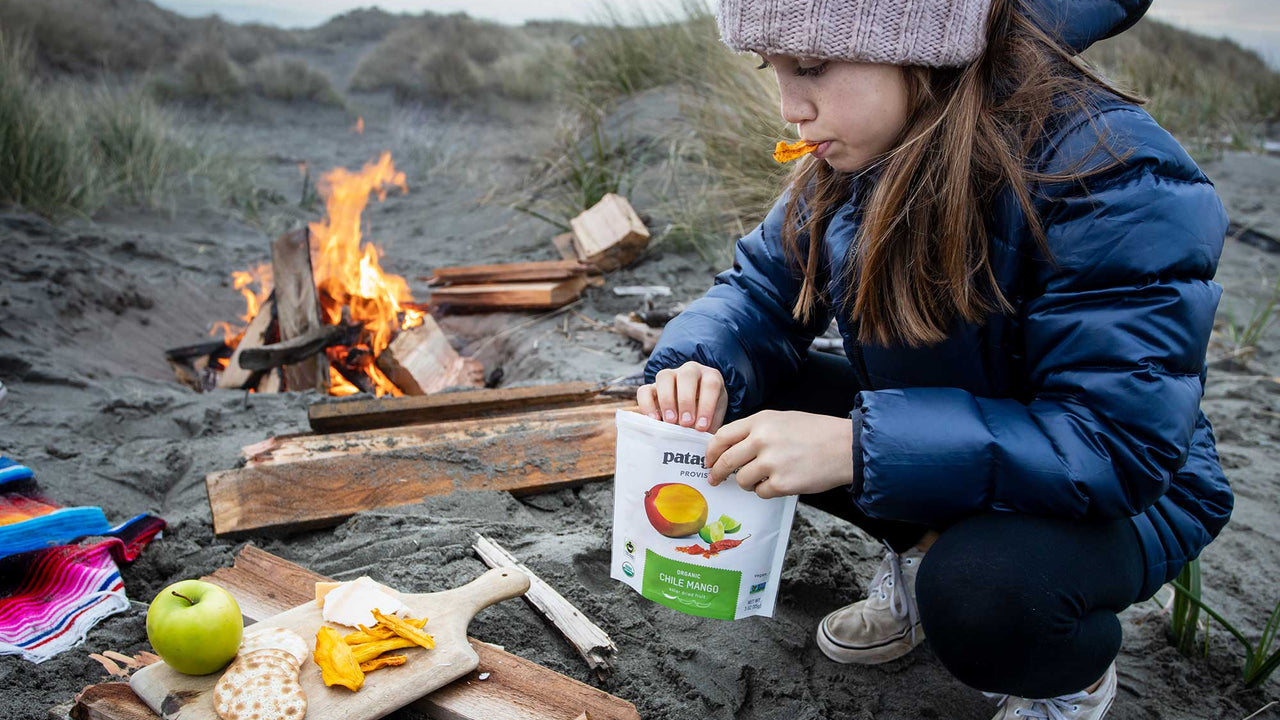 A child at the beach in knitted had and puffy coat snacks on Patagonia Provisions' Chile Mango slices beside a small bonfire