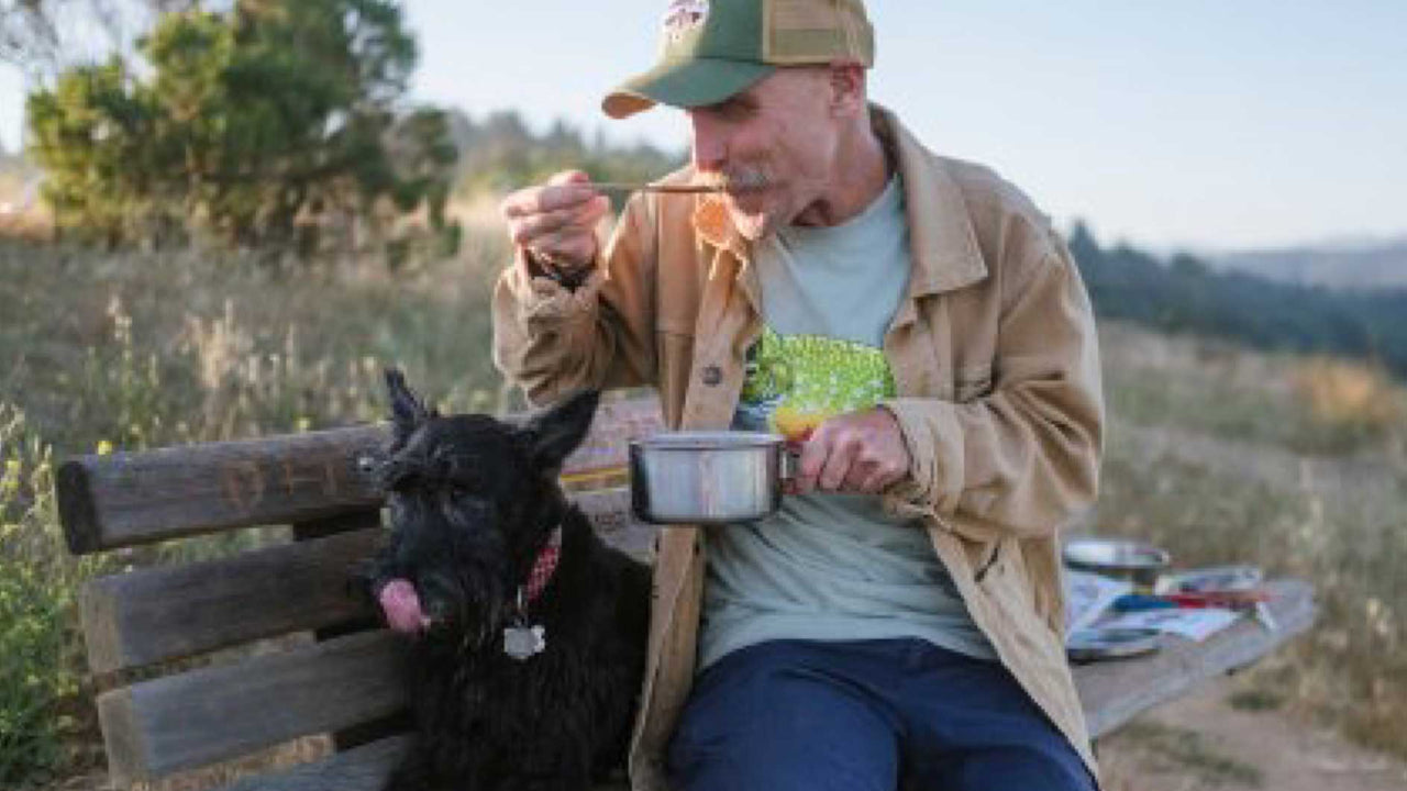 A man wearing baseball cap and jacket sits on a hillside bench beside a Scottie dog and scoops a meal made with Patagonia Provisions foods from a small pot