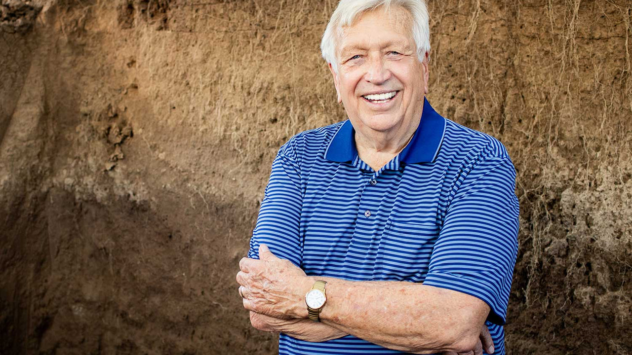 Photo of Wes Jackson smiling with arms crossed in front of a wall of dirt exposing the long roots of the Kernza plant