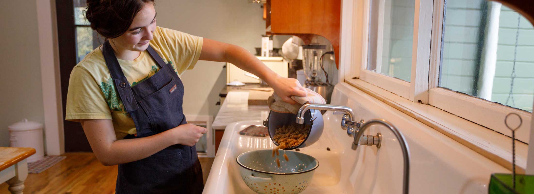 A woman wearing a blue Patagonia apron drains Patagonia Provisions Kernza pasta through a colander in a white enamel sink