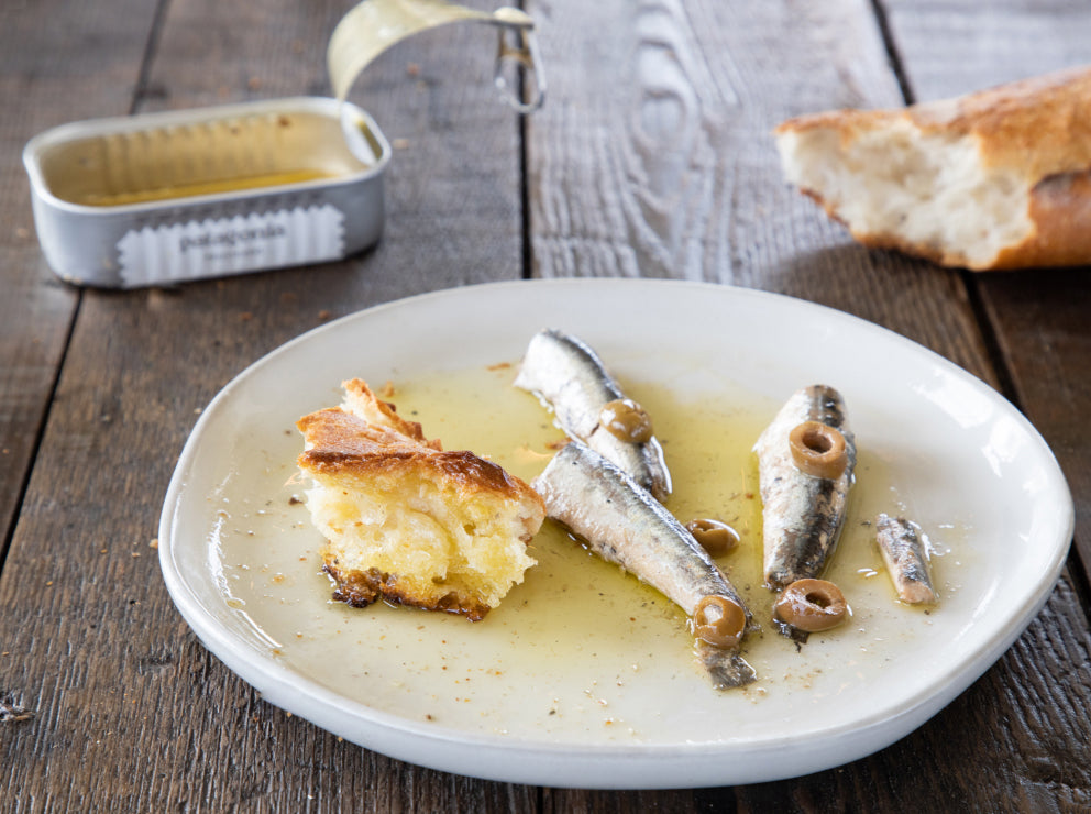 White ceramic plate with Patagonia Provisions Anchovies, olives, and oil, on a wooden table beside crusty bread and open tin
