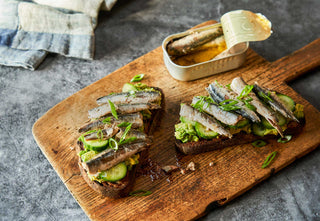 An open tin of Patagonia Provisions Tinned Anchovies on a board beside two open faced toasts topped with avocado and anchovies