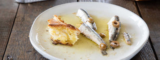A Patagonia Provisions Spanish White Anchovies on a white ceramic plate with olive oil, olives, and a piece of chunky bread, on a wooden table