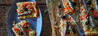 A rustic pizza topped with Patagonia Provisions Anchovies, cartelized onion and red peppers, on top of a wooden board and a blue serving plate