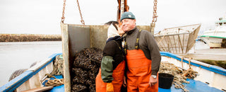A man and woman on a boat wearing protective overalls and hats and the woman is kissing the man on his cheek