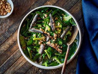 Top down view of Patagonia Provisions Fried White Anchovy, Broccoli and Roasted Almond Salad recipe in a ceramic bowl on wooden surface beside deep blue cloth napkin and small bowl of chopped almonds