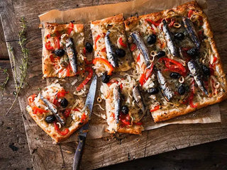 Patagonia Provisions Lemon Olive Spanish White Anchovies top a tart with olives, onions, red pepper and lemon, served on parchment paper on a wooden board