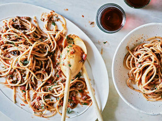 A large serving dish of Spaghetti with Puttanesca Sauce beside a bowl full of pasta and a glass of red wine