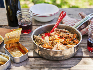 A metal pan filled with Patagonia Provisions Seafood Paella rests on a picnic table beside an open can of mussels, enamel bowls, and jam jar glasses of wine