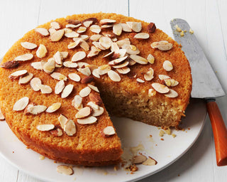 A golden honey, lemon, and olive oil cake topped with honey syrup and sliced almonds, on a plat with cake knife