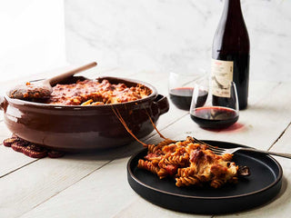 A large ceramic serving bowl and plate of Chorizo Baked Pasta beside glasses and bottle of red wine