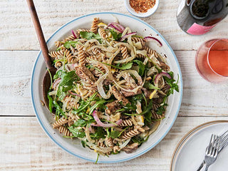 Large white serving bowl filled with Patagonia Provisions Organic Kernza Fusilli pasta mixed with vegetables and Lemon Caper Mackerel, on a white table beside glass and bottle of rosé