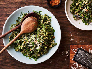 A large white ceramic serving bowl filled with dark green pesto and fusilli sits on a dark wooden table beside a cheese grater with parmesan