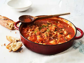 A large red enamel pot filled with Seafood Stew rests on a white tablecloth beside fresh pieces of baguette