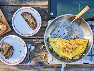 A folded-in-half omelet stuffed with Patagonia Provisions Wild Salmon and colorful vegetables in a skillet on a camp stove, beside two plates with slices of toast