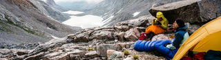 Backpackers sit in sleeping bags on a rocky mountain, having a meal
