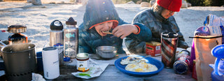 Winter camping family shares a meal at a picnic table with Patagonia Provisions foods and drink containers