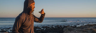 A man in a warm hooded jacket stands on the beach in front of the sea eating a snack with Patagonia Provisions savory seeds