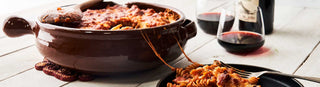 A large ceramic pot of warm cooked pasta on a white wooden table beside a plate of pasta and bottle with two glasses of red wine