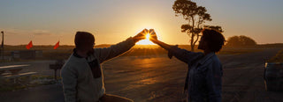 Two people in silhouette join their raised hands to form a heart around the setting sun