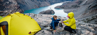 An adult and child prepare a meal outside a bright yellow tent on a rocky outcropping looking over a river