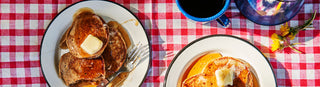 Tin plates with fresh pancakes sit on a red and white checked tablecloth next to blue enamel coffee cup and dandelions
