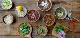 Looking down at a wooden table covered with bowls of Patagonia Provisions soups and chilis and fresh topping ingredients