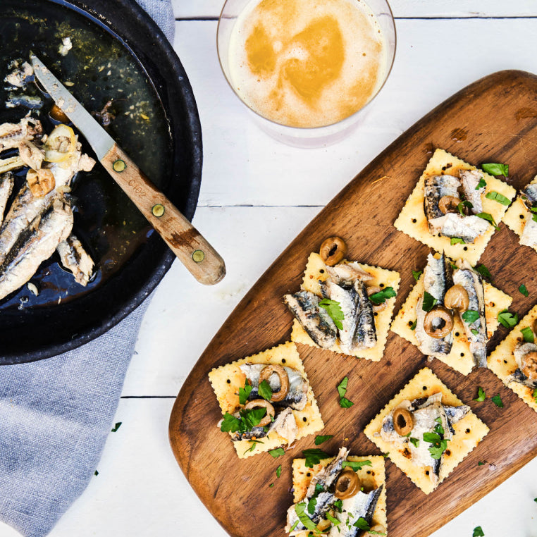 A wooden serving board covered with saltine crackers topped with olives and Patagonia Provisions Spanish White Anchovies rests on a white table beside a skillet with anchovies