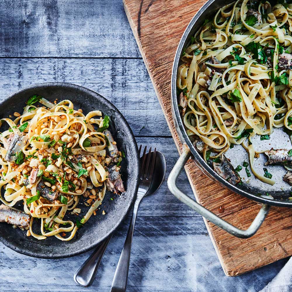 A pan and dark ceramic bowl of Patagonia Provisions Spanish White Anchovies and linguine with toasted breadcrumbs