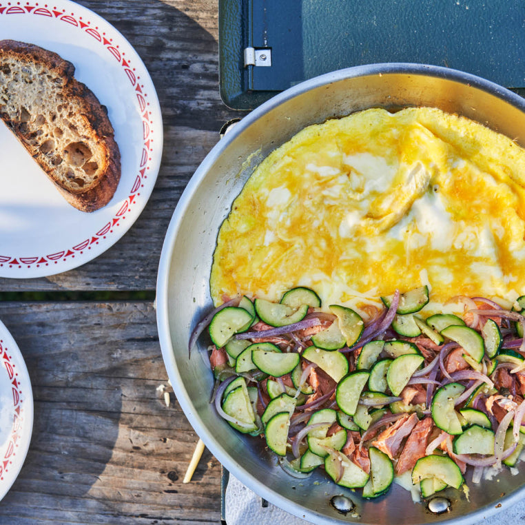 Campsite cooking shot of large metal skillet half filled with a salmon and zucchini omelette about to be folded
