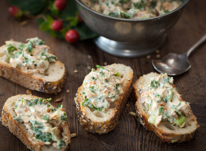 Slices of seeded baguette on a wooden surface are topped with dip made with Patagonia Provisions Smoked Salmon