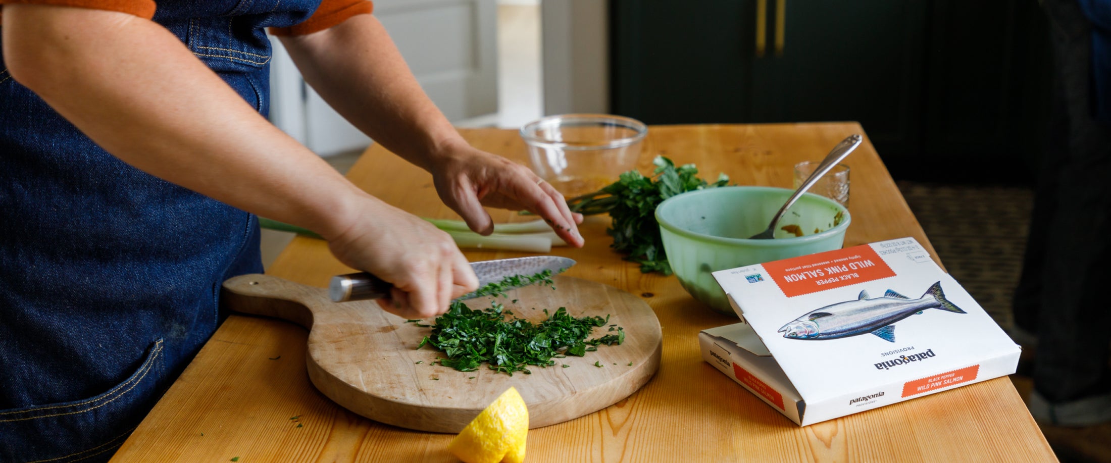 A home chef chops herbs at a wooden kitchen counter, beside a box of Patagonia Provisions Wild Pink Salmon