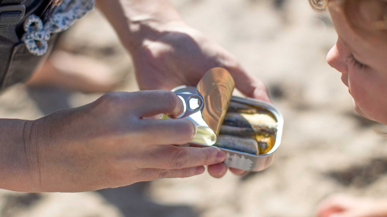 An adult opens a can of Patagonia Provisions Anchovies for a child on a beach