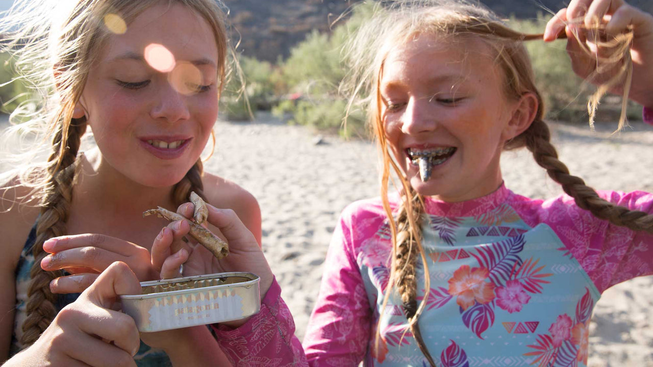 Two young friends in swimsuits share Patagonia Provisions Mackerel from a tin on a beach
