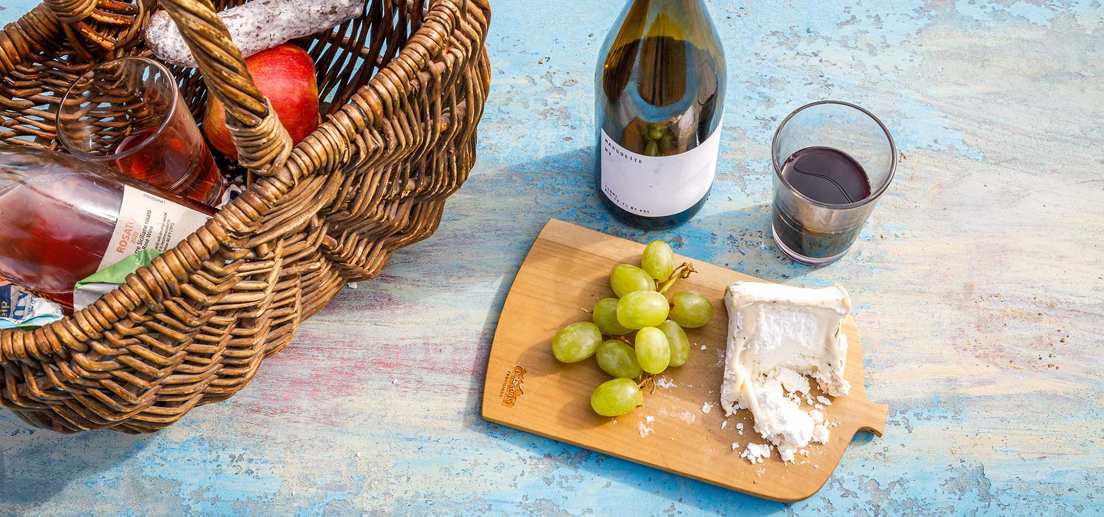A picnic basket rests on a pale blue table outdoors, beside a board of grapes and cheese and a bottle of Patagonia Provisions red wine
