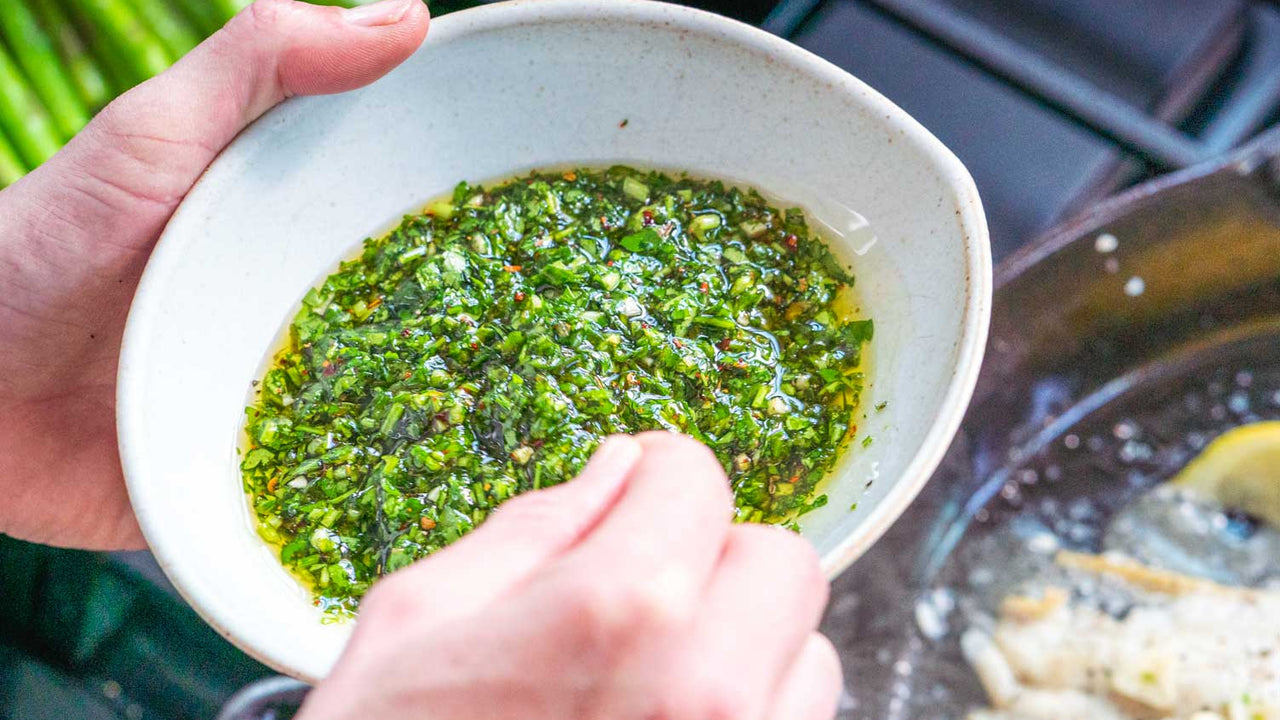 A white bowl of fresh chimichurri sauce, held over a skillet of fish and lemon