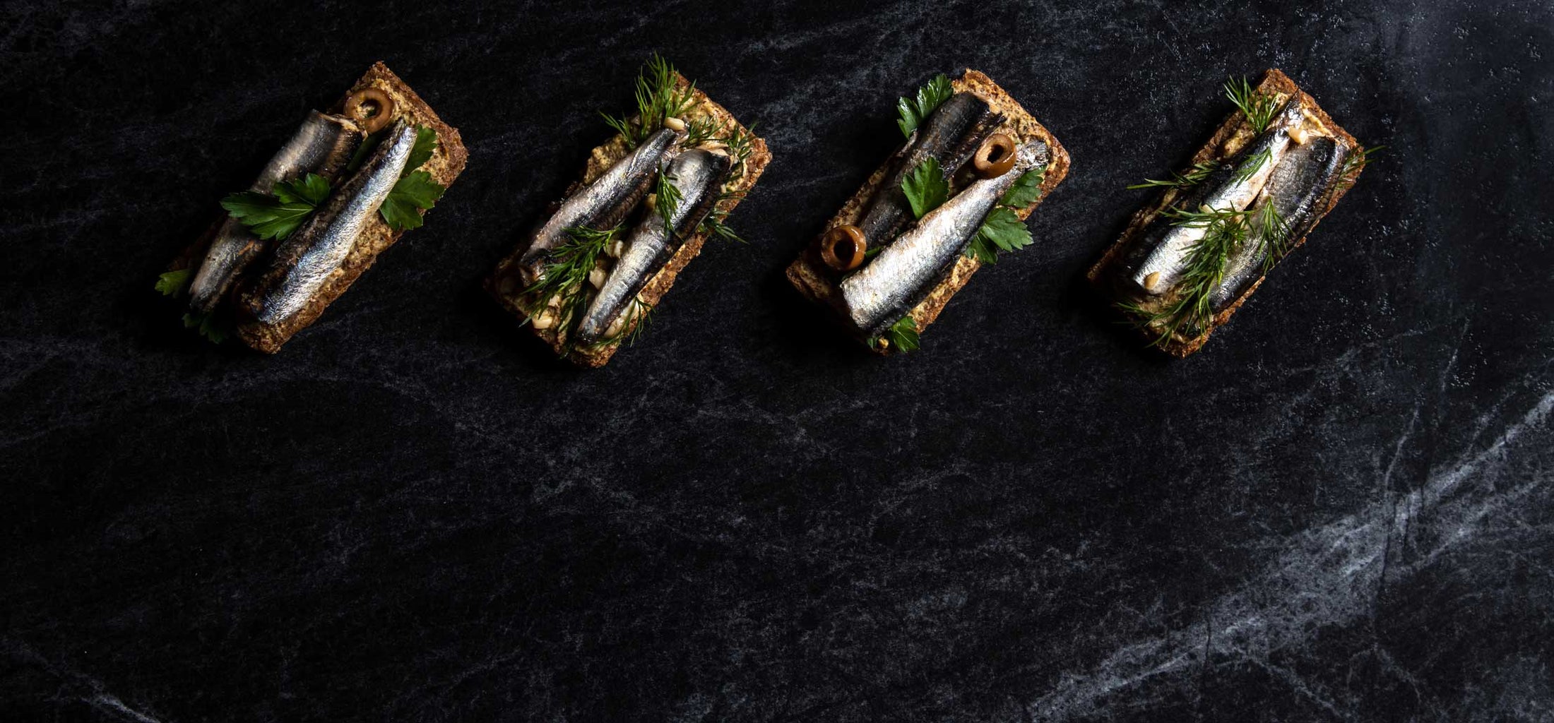 A row of toasts topped with slices of Spanish White Anchovies, herbs, and olives, on a dark stone tabletop