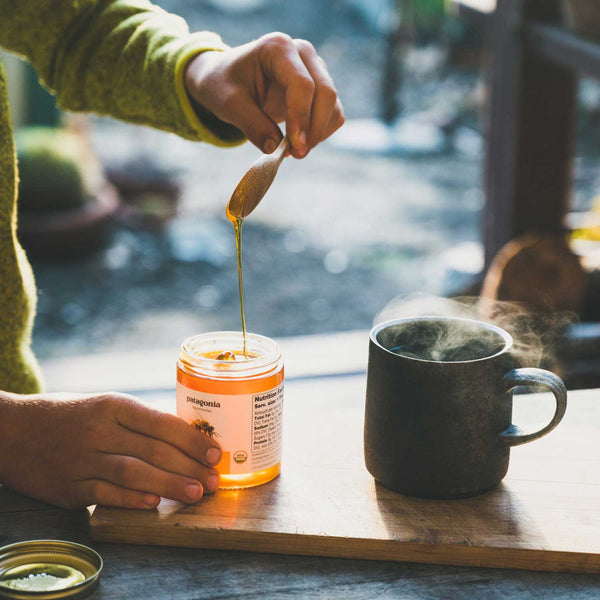 Close shot of hands scooping Patagonia Provisions Organic Moloka'i Honey from the jar with a small wooden spoon, to drizzle into a steaming ceramic cup