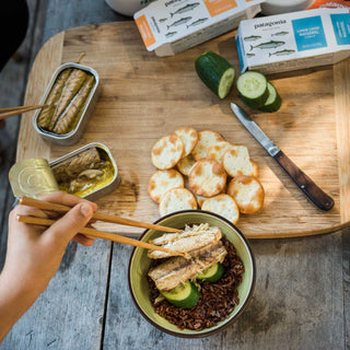 Hands use chopsticks to add pieces of Patagonia Provisions Mackerel from open tins to a bowl of dark rice and cucumber, next to a cutting board with crackers and sliced cucumber