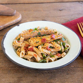 Long pieces of Patagonia Provisions Spanish Paprika Mackerel top a plate of fresh pasta, tomato, and herbs, in white enamel dish on a wooden table