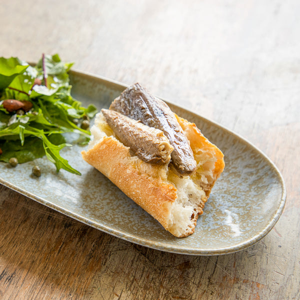 Oblong ceramic dish with salad and chunk of baguette topped with Patagonia Provisions Smoked Mackerel on top of a wooden table