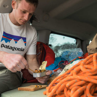 A climber wearing a Patagonia tshirt sits in a van filled with climbing gear, opening a can of Patagonia Provisions' Savory Sofrito Mussels and scooping them out onto a piece of wheat bread